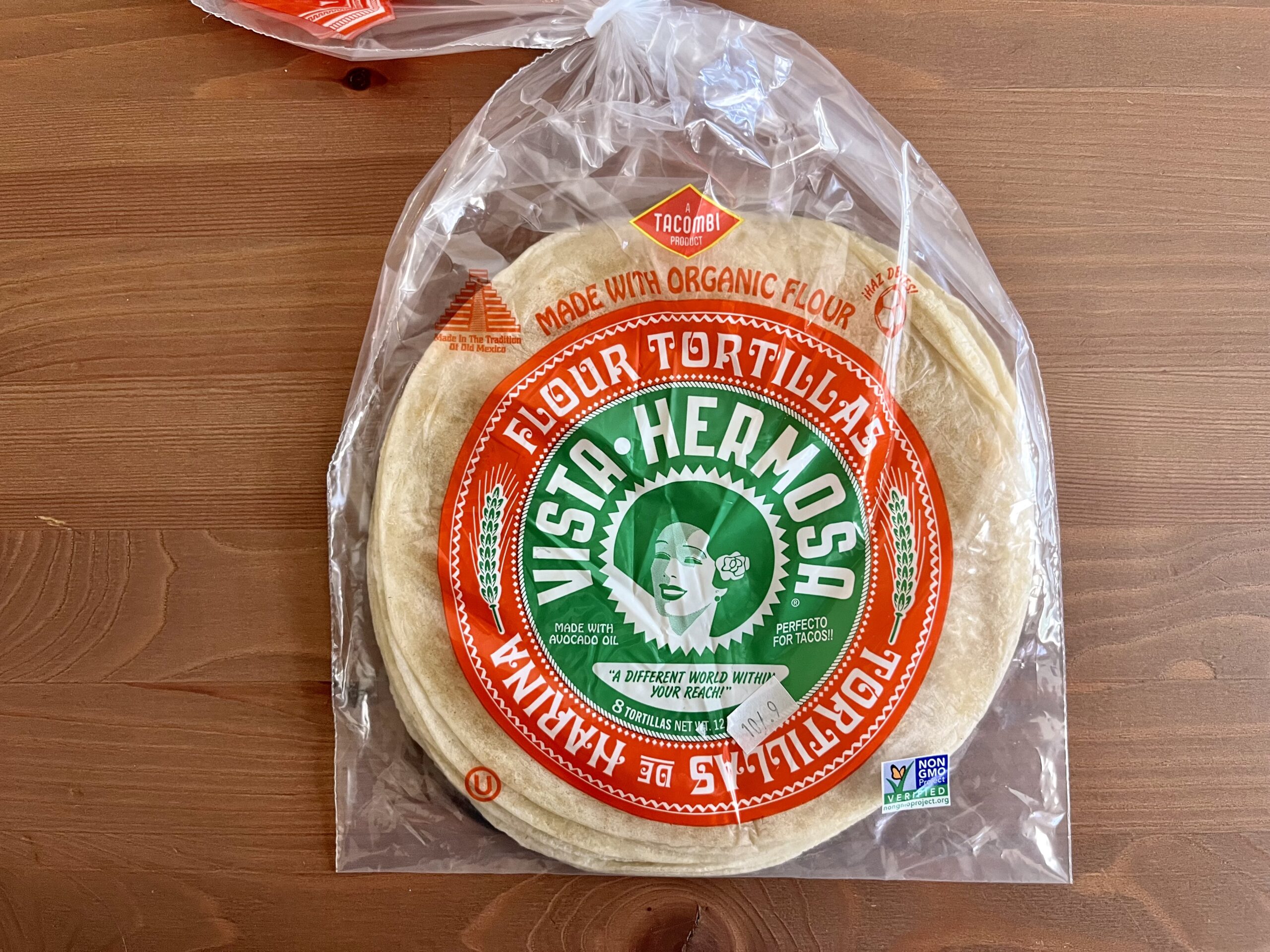 A package of Vista Hermosa flour tortillas made with organic flour sits on a wooden surface. The clear bag features a green and red label with a white graphic of a woman's face and text describing the tortillas.