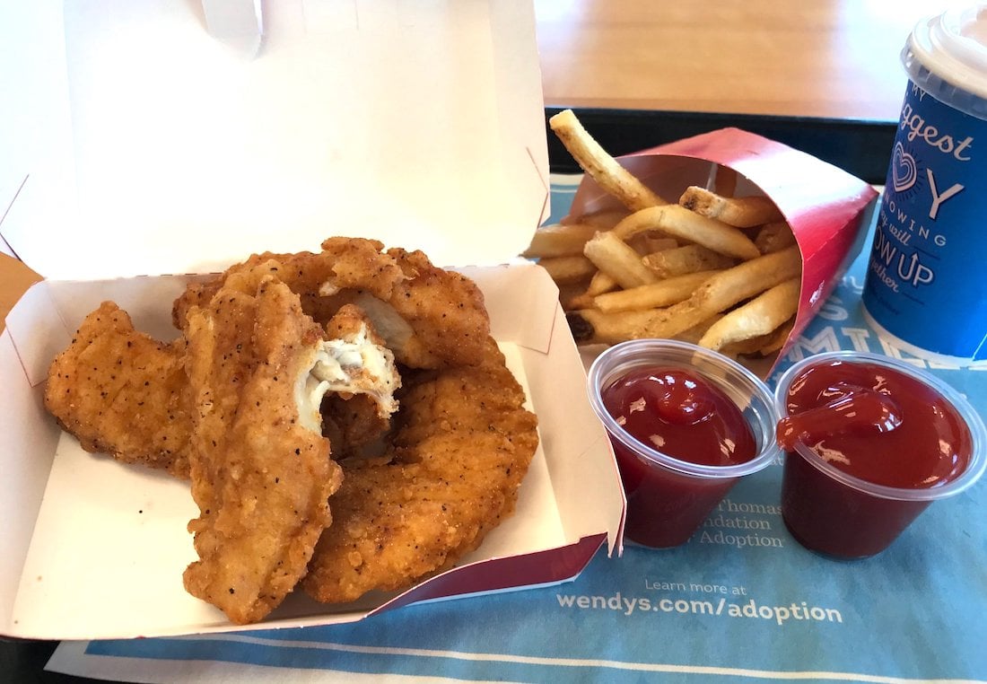 A meal with crispy chicken tenders in a white box, a red container of French fries, two cups of ketchup, and a blue soft drink cup on a tray with a promotional paper mat.