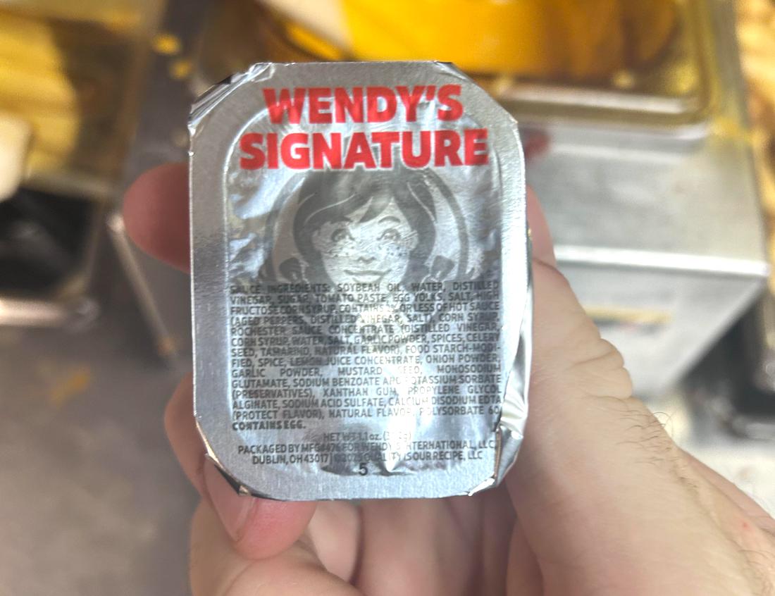 A hand holds a container of Wendy’s Signature sauce with a silver foil lid displaying the brand’s logo and ingredient list in small text. The background is blurred and shows food preparation equipment.