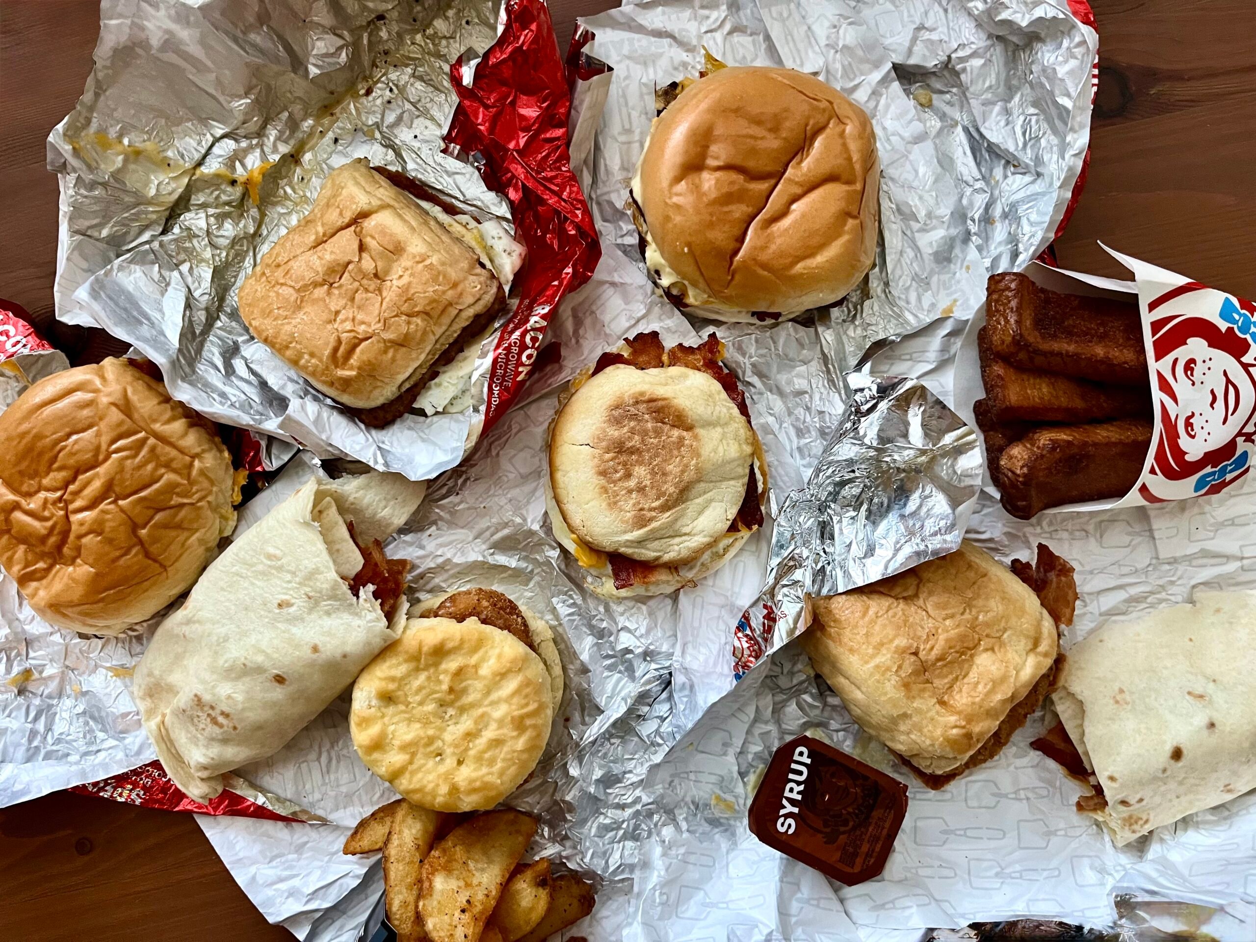 An assortment of fast food breakfast items, including several wrapped sandwiches, biscuits, potato wedges, burritos, packets of syrup, and a container of French toast sticks, laid out on a wooden table.