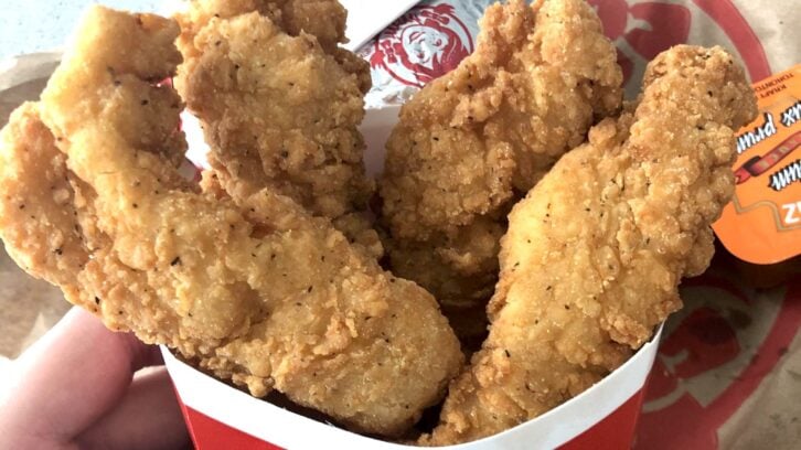 A hand holding a container of crispy, golden-brown chicken tenders from Wendy's, with a cup of dipping sauce visible in the background.