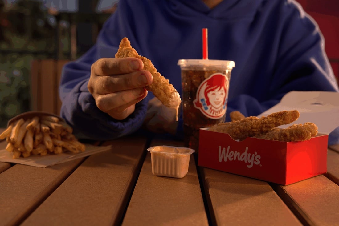 A person in a blue hoodie dips a Wendy's chicken strip into sauce at an outdoor table, enjoying fries, a drink, and a red Wendy's box—savoring a classic meal at this new fast-food spot.