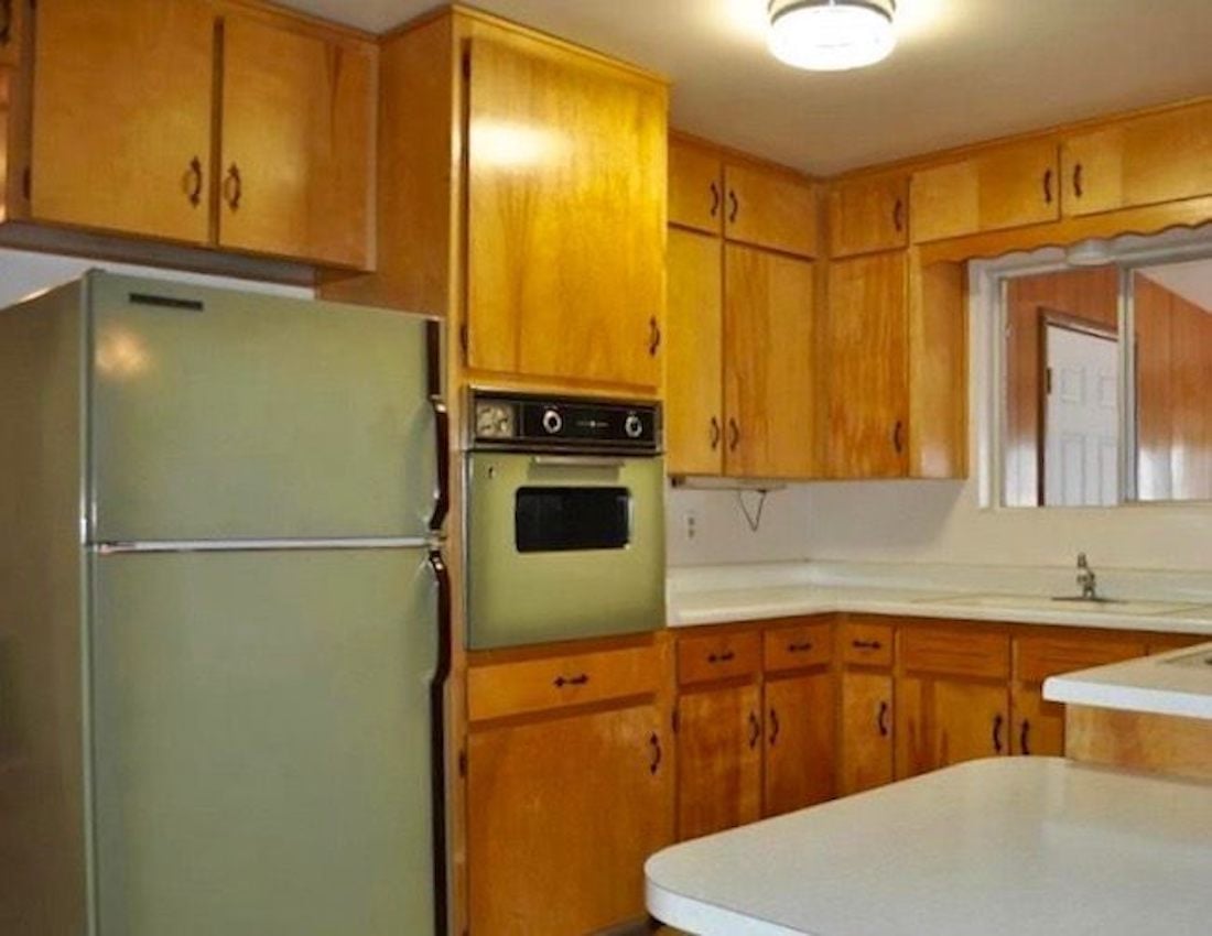 A vintage kitchen with honey-colored wooden cabinets, a green refrigerator, a built-in oven, white countertops, and a small sink under a window. The lighting is bright, and the decor appears retro.
