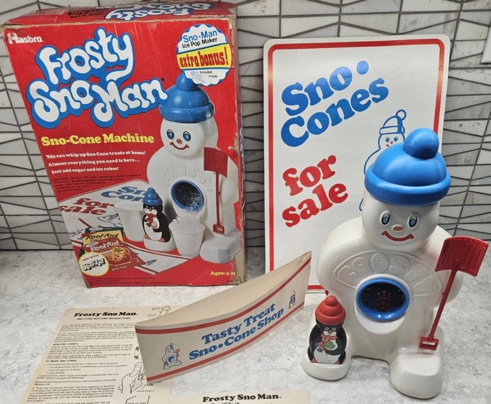 A vintage Frosty Sno Man Sno-Cone Machine toy with its original box, instruction sheet, snowman-shaped machine, shovel, flavor bottle, and a sign reading "Sno Cones for sale" displayed on a countertop.