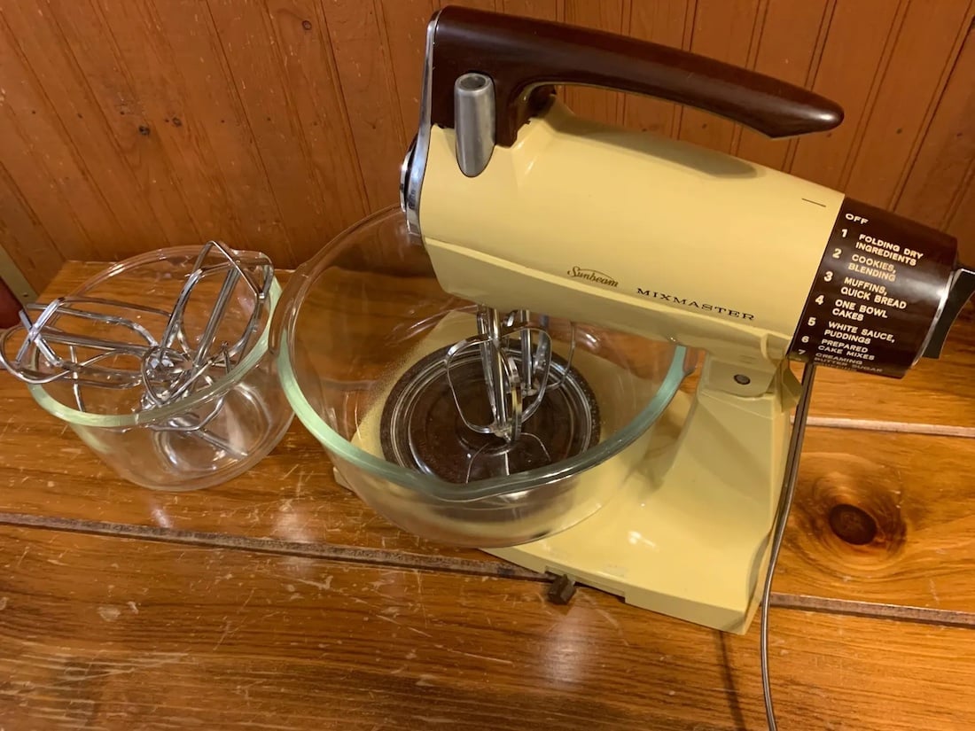 A vintage yellow electric stand mixer with a glass mixing bowl and metal beaters sits on a wooden countertop. A second glass bowl and extra beaters are beside it. The mixer's speed settings are visible on a brown dial.