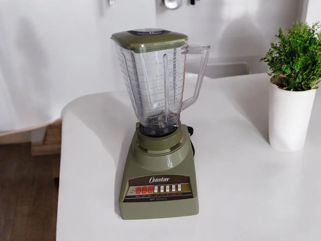 A vintage Osterizer blender with a green base and clear pitcher sits on a white countertop next to a potted plant. The blender has several buttons labeled with different speed settings.