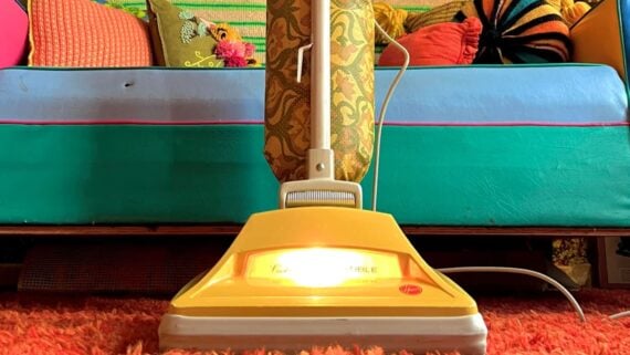A vintage upright vacuum cleaner with a yellow base and patterned cloth bag stands on an orange shag carpet in front of a teal sofa with colorful patterned pillows.