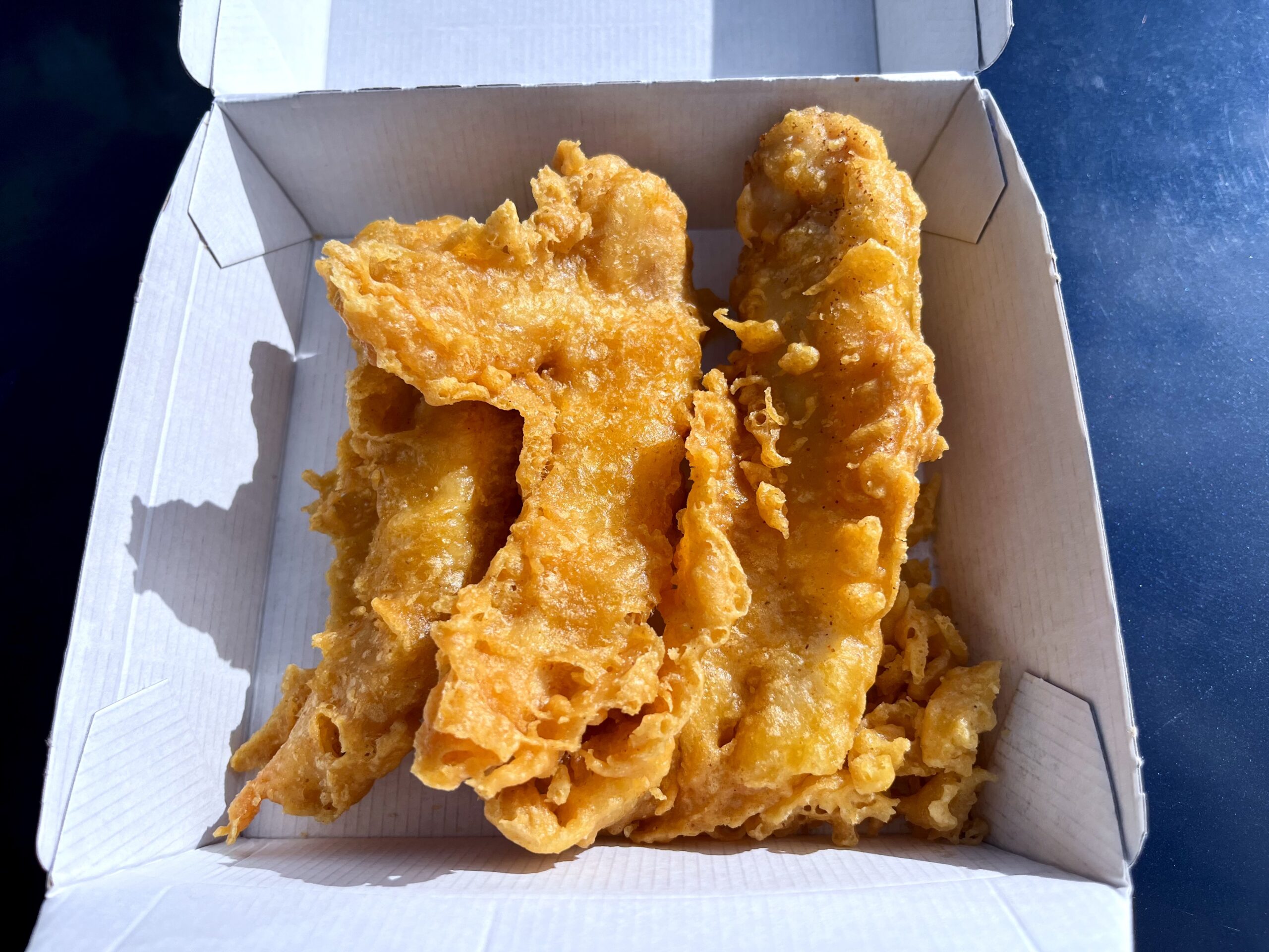 Two pieces of golden, crispy battered fish placed side by side in a white cardboard takeout box, photographed in bright natural light.