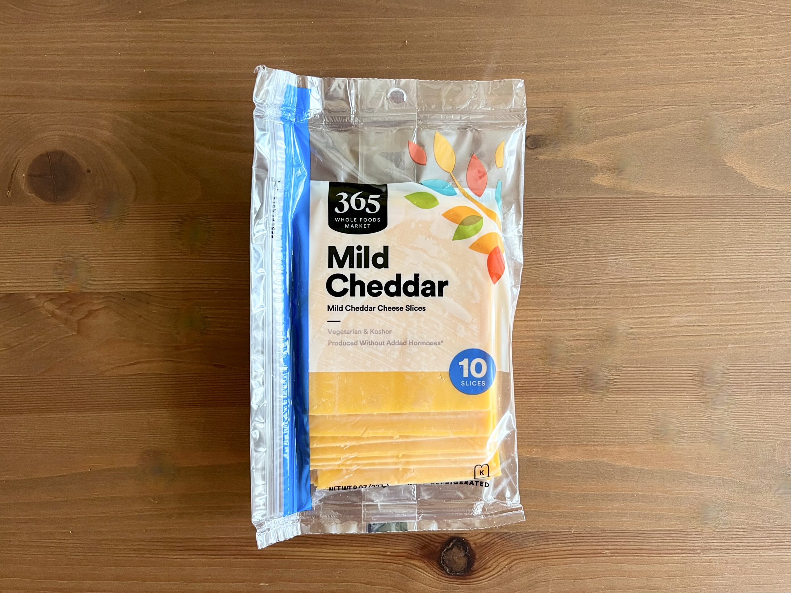 A package of 365 Whole Foods Market Mild Cheddar cheese slices sits on a wooden surface. The clear plastic packaging shows yellow cheese slices inside and a label indicating there are 10 slices.