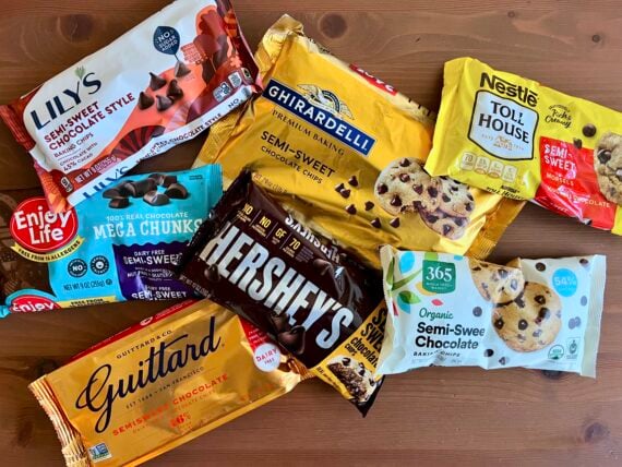 Assorted bags of semi-sweet chocolate chips from brands like Lily’s, Ghirardelli, Nestlé Toll House, Enjoy Life, Hershey’s, Guittard, and 365, displayed on a wooden surface.