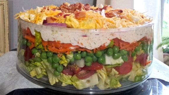 A clear glass bowl is filled with layered salad, including lettuce, green peas, tomatoes, onions, shredded carrots, a thick layer of creamy dressing, shredded cheese, chopped eggs, and bacon bits on top.