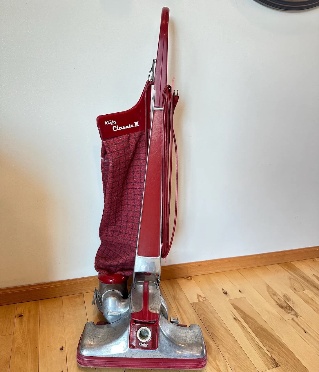 A vintage red and silver Kirby Classic III vacuum cleaner stands on a wooden floor against a white wall. The vacuum has a cloth bag and a power cord wrapped around the handle.