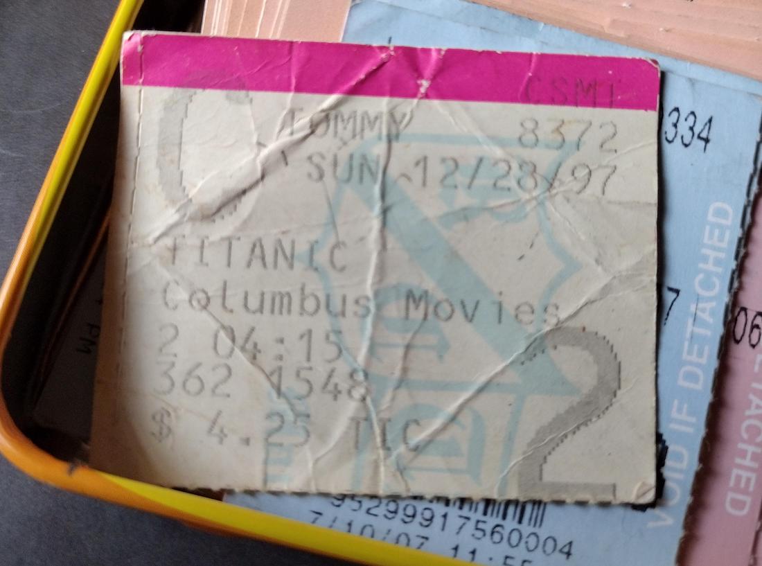 A worn movie ticket stub for "Titanic" at Columbus Movies dated Sunday, 12/28/97, with a showtime of 2:15. The ticket price is $4.25, and it's placed on top of other stubs in a container.
