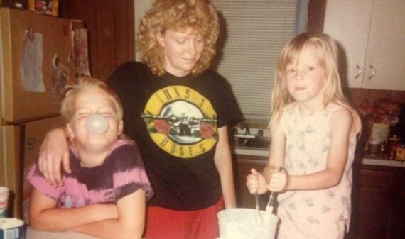 A woman with curly blonde hair stands in a kitchen with two children. One child blows a bubblegum bubble, and the other stirs a bowl. The woman wears a Guns N' Roses t-shirt and smiles at the children.