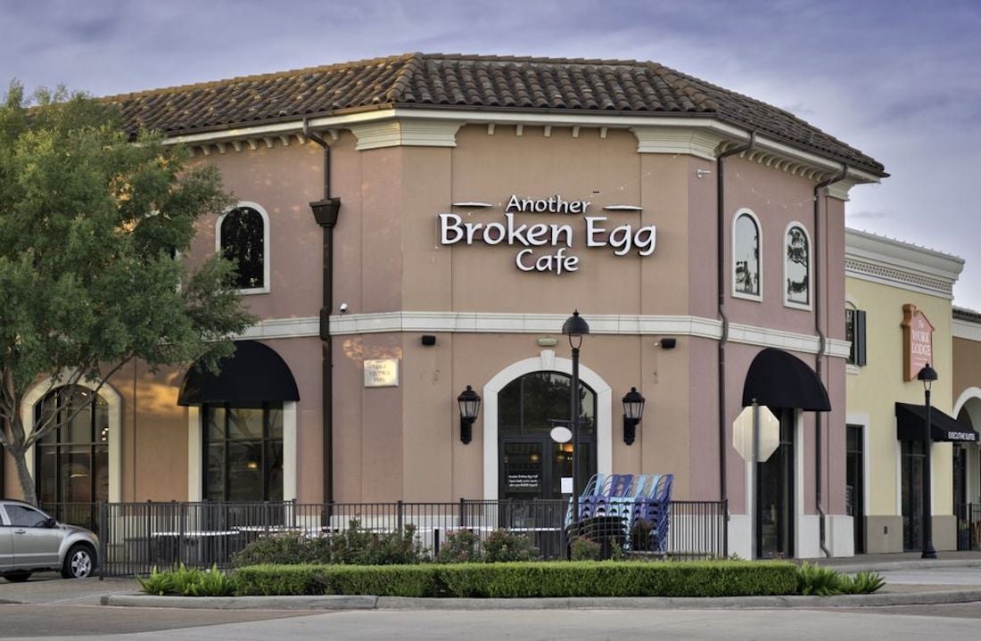 A tan, two-story building with a sign reading "Another Broken Egg Cafe" above the entrance, large windows, black awnings, and a small fenced patio out front. A tree and parked car are visible beside the cafe.
