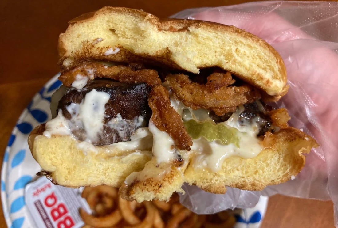 A close-up of a hand holding a half-eaten cheeseburger with fried onions, pickles, and creamy sauce on a soft bun. Curly fries are visible on a paper plate in the background.