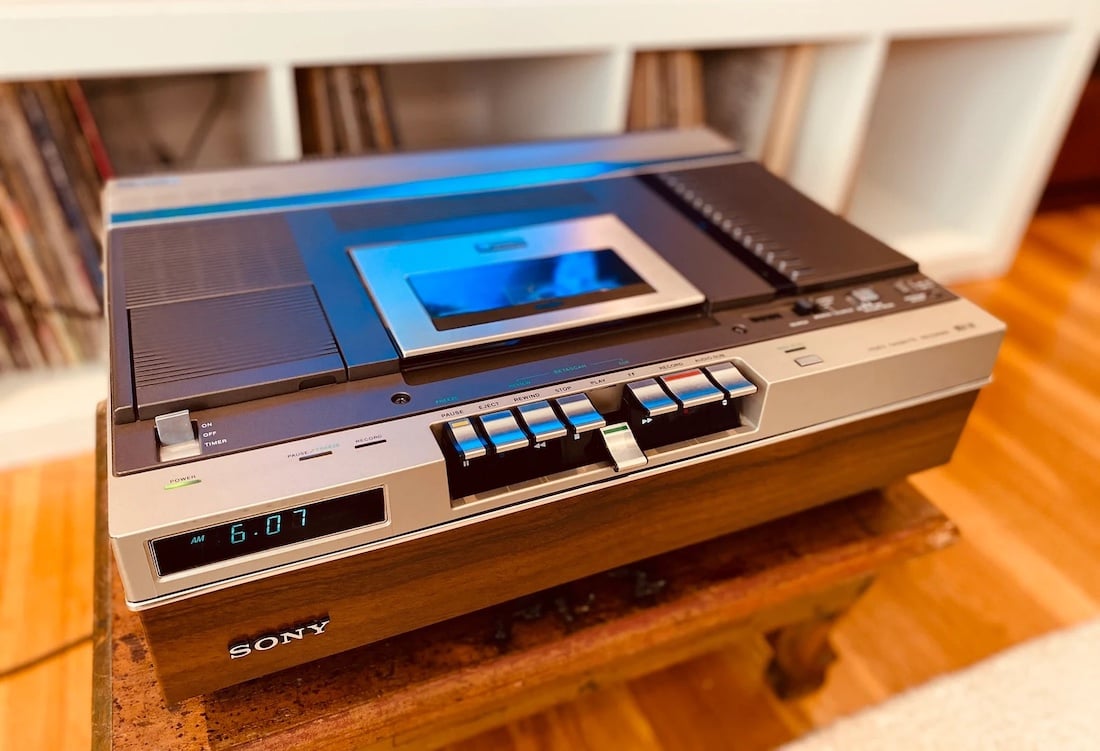 A vintage Sony Betamax VCR sits on a wooden table, displaying the time “6:07” on its digital screen. Shelves with vinyl records are visible in the background.