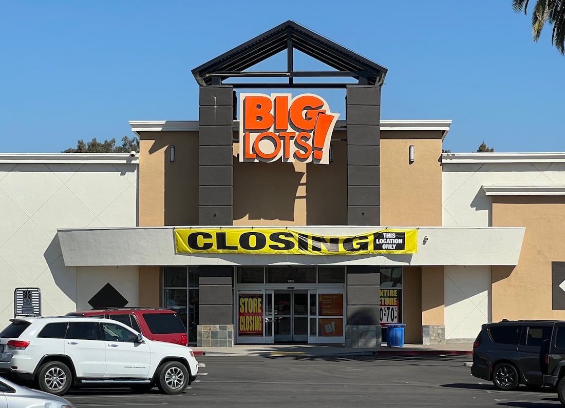 The front of a Big Lots! store with a large yellow "CLOSING!" banner above the entrance, several cars parked in front, and a clear blue sky overhead.