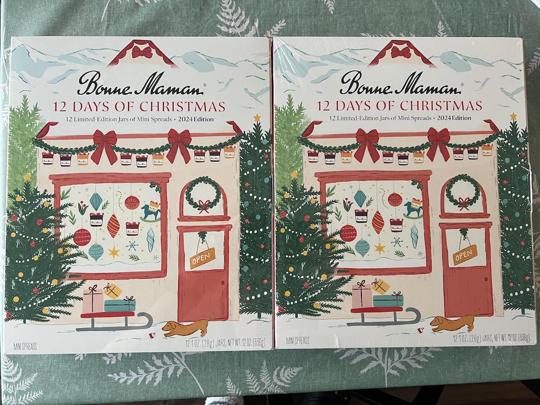 Two boxes of Bonne Maman 12 Days of Christmas jam gift sets, featuring a festive illustration of a shop decorated with holiday garlands, a wreath, and gifts, set against a snowy background with Christmas trees.