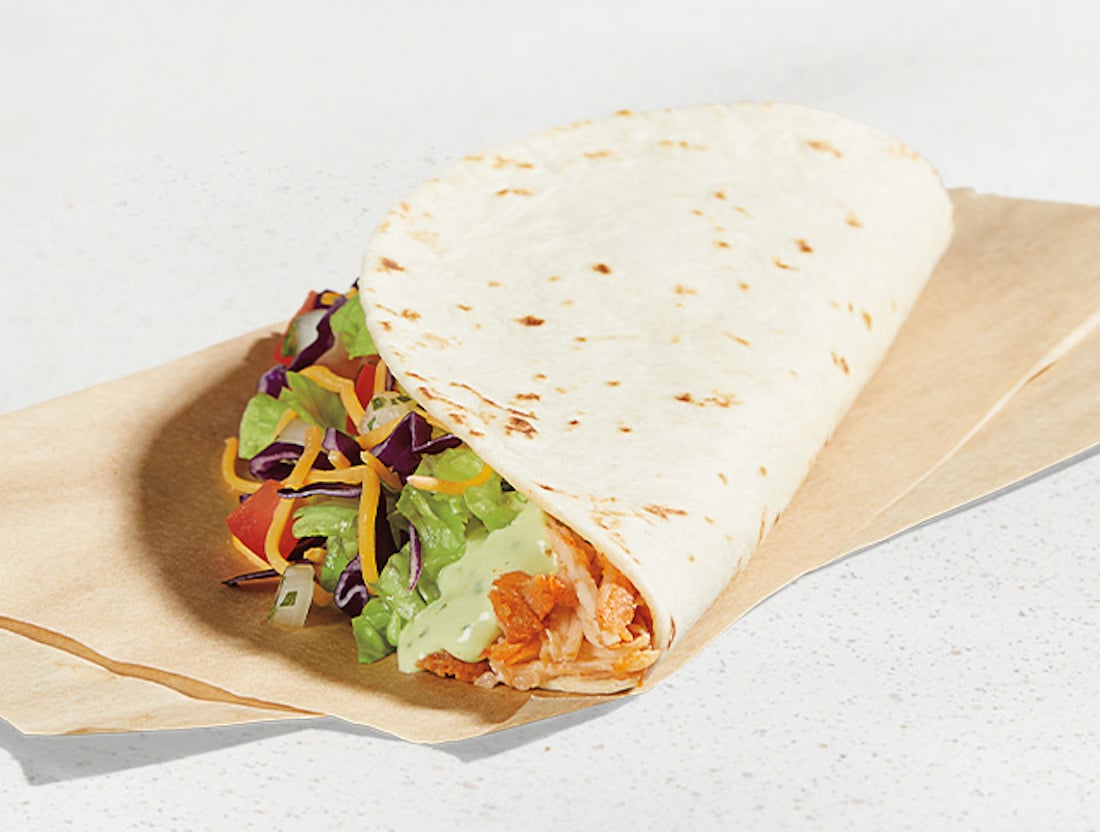 A soft flour taco filled with shredded chicken, lettuce, cheese, diced tomatoes, and a creamy sauce, partially wrapped in brown paper on a light surface.