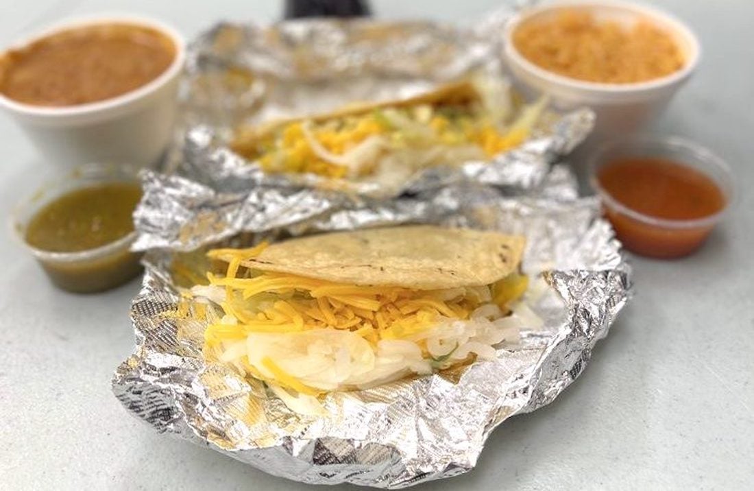 Two corn tacos filled with lettuce, onions, and shredded cheese sit on foil, accompanied by small cups of salsa, refried beans, and Mexican rice in foam containers on a light-colored table.
