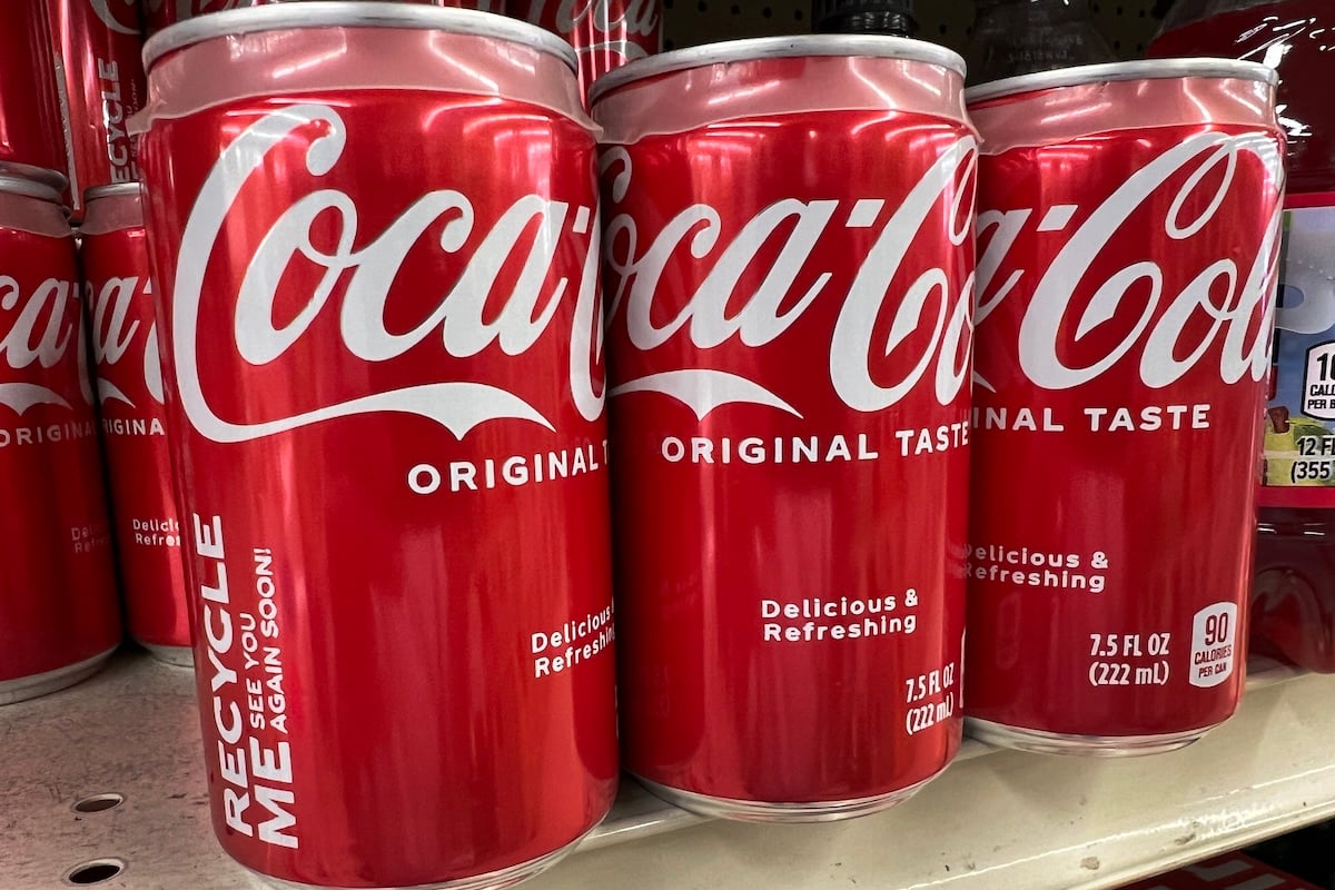 Three red Coca-Cola cans labeled "Original Taste" and "Delicious & Refreshing" are lined up on a store shelf. Each can has a volume of 7.5 fl oz (222 mL) and a "Recycle Me" message printed on the side.