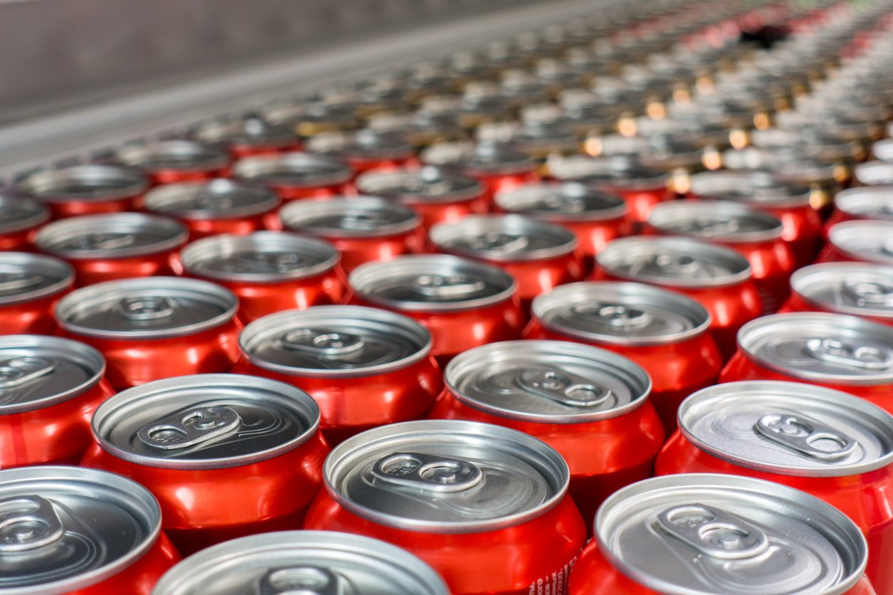 Rows of unopened red aluminum cans, likely soda, are arranged closely together on a production line in a factory setting.