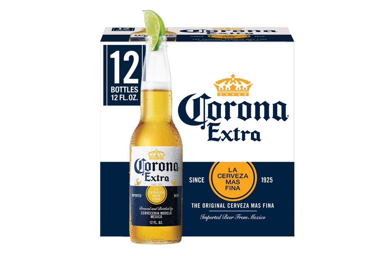 A bottle of Corona Extra beer with a lime wedge in the neck stands in front of a 12-pack box. The box displays the Corona Extra logo and reads "La Cerveza Mas Fina" with details about the beer's origin from Mexico.