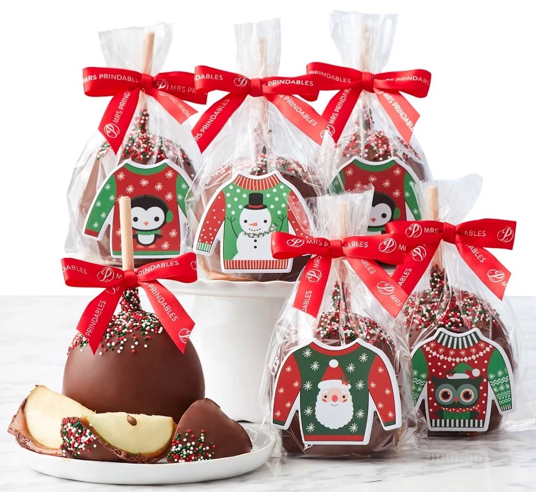 Six chocolate-covered caramel apples decorated with festive Christmas sweater designs, each wrapped in clear plastic and tied with red ribbons. One apple is sliced to show caramel and apple inside.