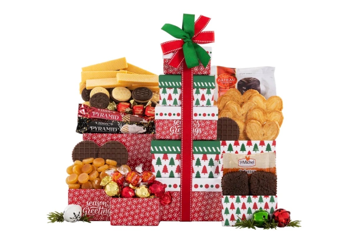A festive holiday gift tower with red, white, and green boxes decorated with Christmas trees and topped with a green and red bow. The boxes are filled with assorted cookies, chocolates, wafers, and candies.