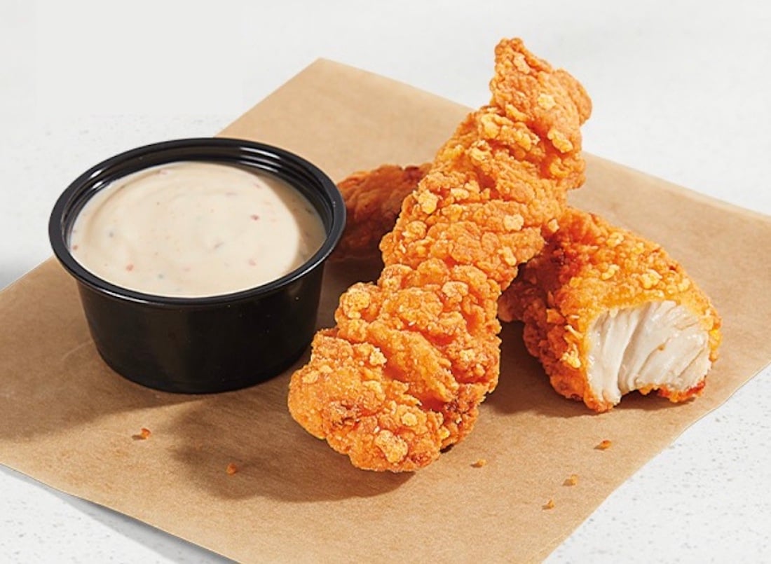 Two crispy fried chicken tenders on brown parchment paper beside a small black cup of creamy dipping sauce, with one tender partially broken to show the white meat inside.