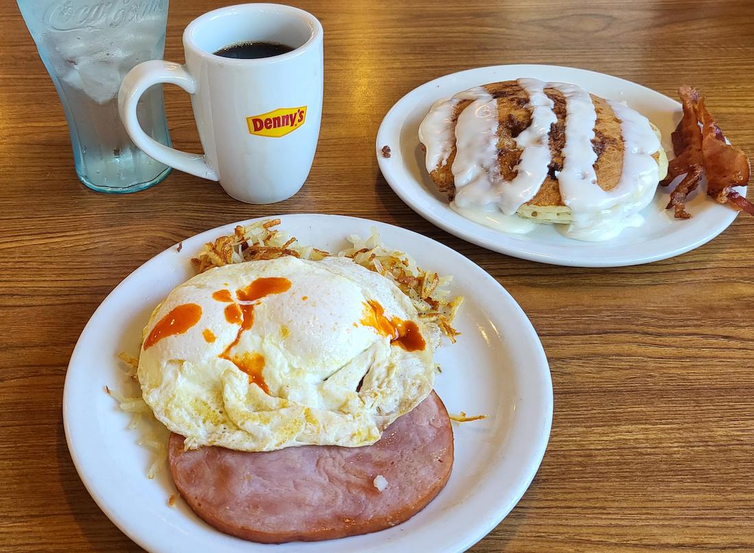 A breakfast meal on a wooden table with ham and eggs over hashbrowns, a side plate of pancakes with white icing and bacon, a cup of coffee in a Denny's mug, and a glass of water.