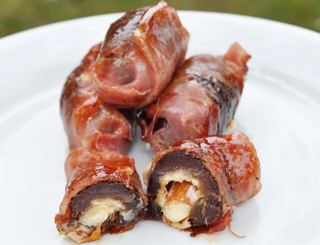 Four bacon-wrapped dates stuffed with cheese are arranged on a white plate, with one piece cut open to show the creamy filling inside. The bacon is cooked to a glossy, slightly crispy finish.