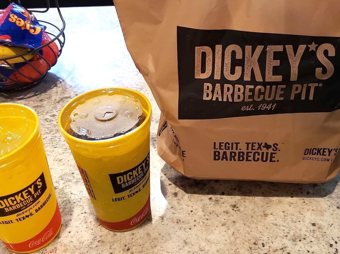 Two yellow Dickey’s Barbecue Pit cups with lids and a large brown Dickey’s Barbecue Pit takeout bag sit on a countertop. A fruit bowl holding apples and lemons is in the background.