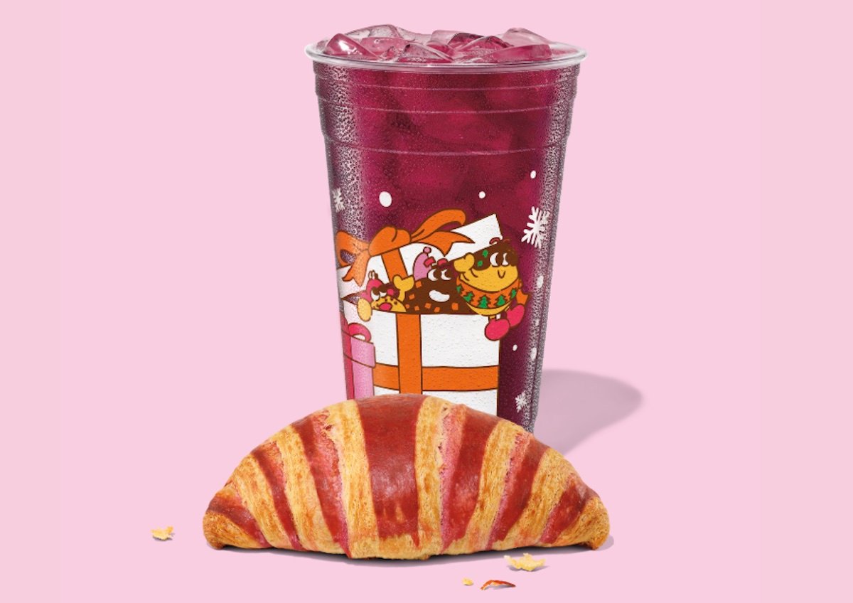 A clear cup of iced purple drink with cartoon gift box design sits behind a golden brown croissant, all set against a light pink background.