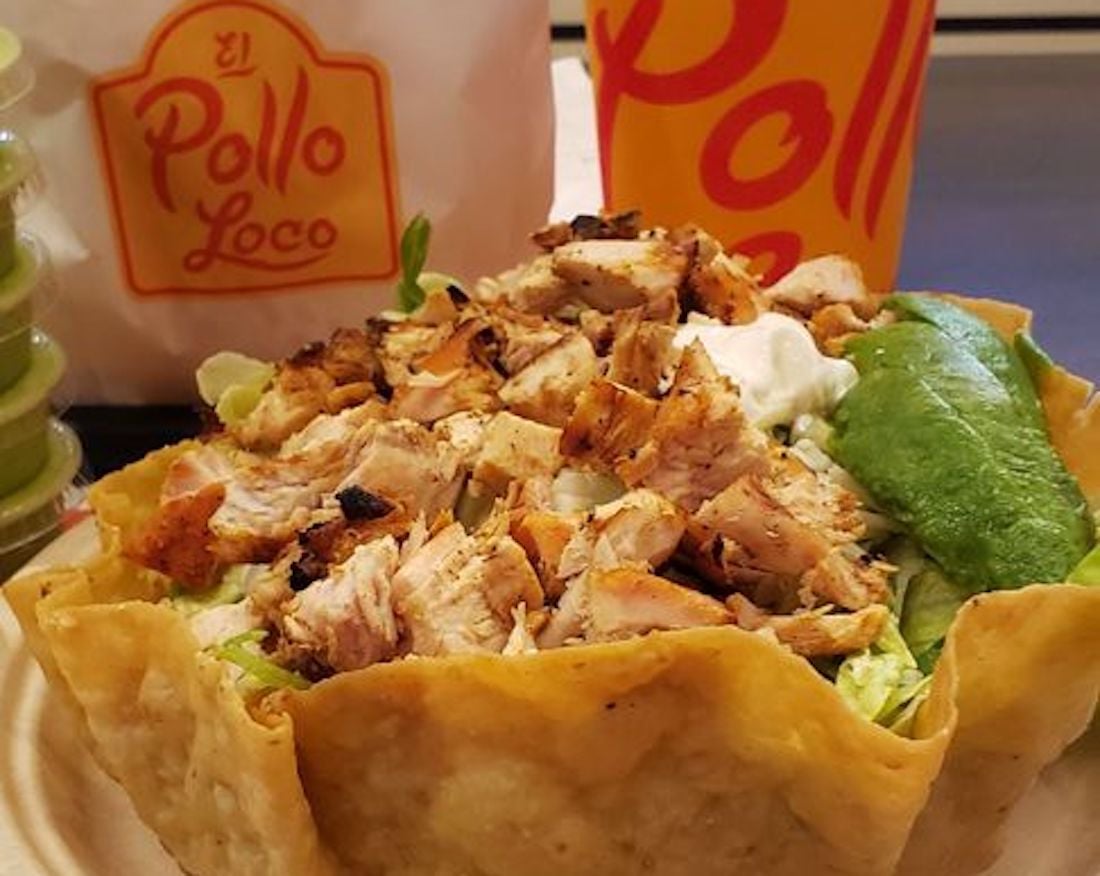 A crispy tortilla bowl filled with grilled chicken, lettuce, sour cream, and avocado sits in front of El Pollo Loco branded bags and a drink cup. Green sauce containers are visible on the side.