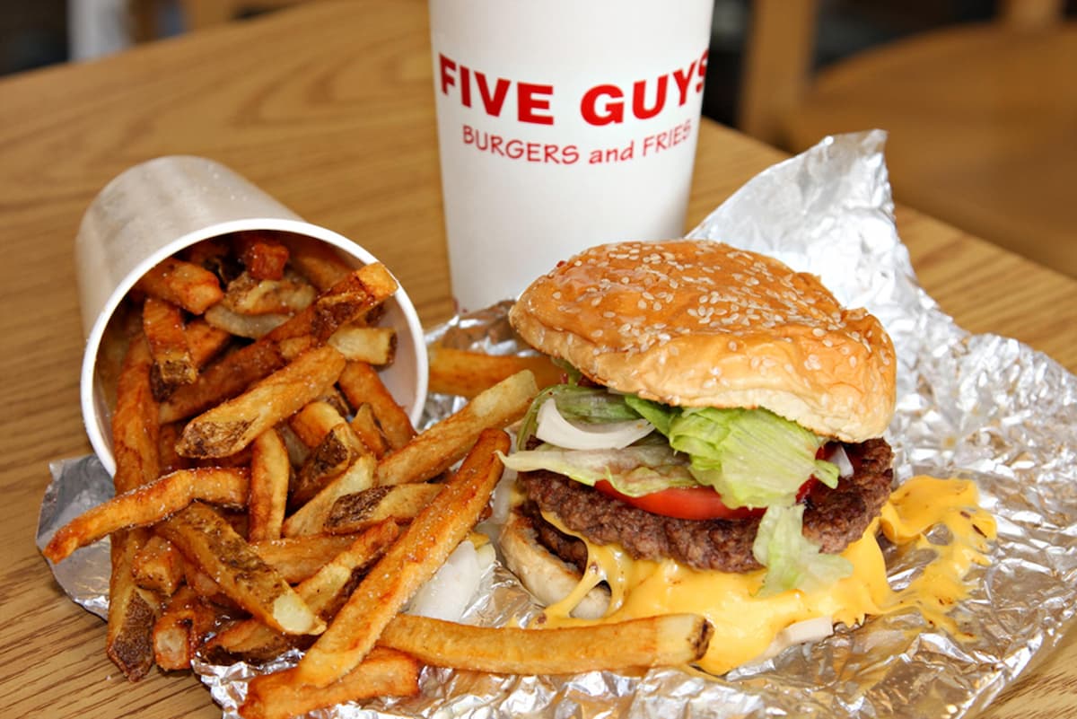 A Five Guys meal with a cheeseburger topped with lettuce, tomato, and onion on foil, fresh-cut fries spilled onto the table, and a large drink—classic fast food from one of the few fast-food chains that never use a microwave.