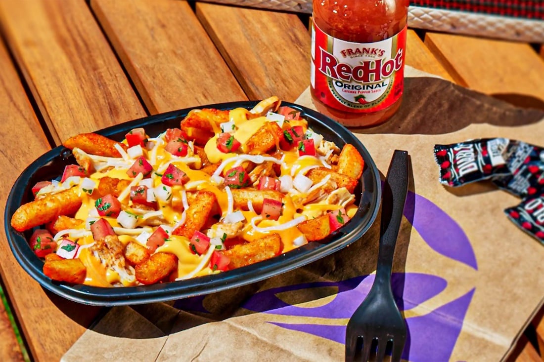 A black tray of loaded nacho fries topped with melted cheese, diced tomatoes, and other toppings sits on a Taco Bell wrapper next to a bottle of Frank's RedHot sauce, hot sauce packets, and a black fork on a wooden table.