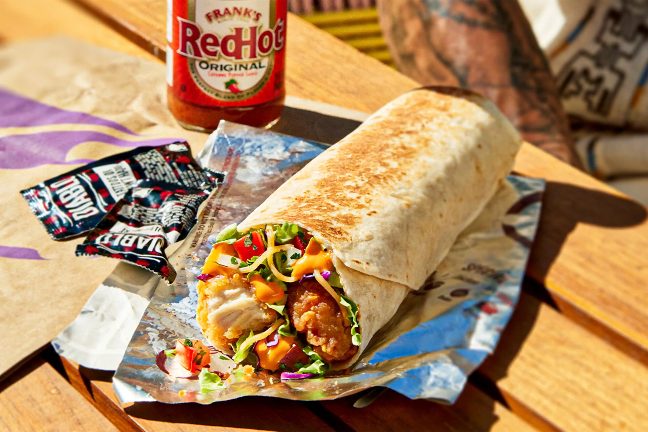 A grilled wrap filled with fried chicken, lettuce, and shredded vegetables sits on foil next to two sauce packets and a bottle of Frank’s RedHot sauce on a wooden table. Part of a tattooed arm is visible in the background.