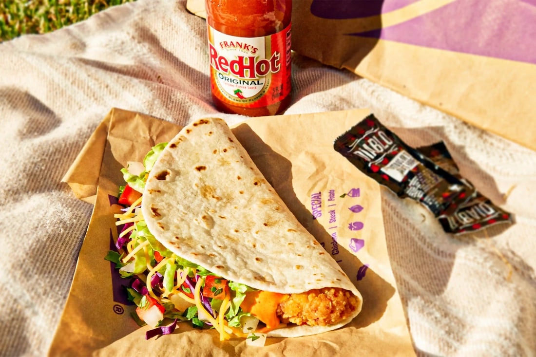 A soft taco filled with fried chicken, shredded cheese, lettuce, and purple cabbage rests on a wrapper. Nearby are packets of hot sauce and a bottle of Frank's RedHot sauce on a blanket.