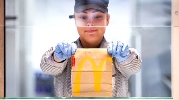 A McDonald's employee wearing a uniform, cap, and blue gloves hands a brown paper bag with a yellow McDonald's logo to a customer across a counter.