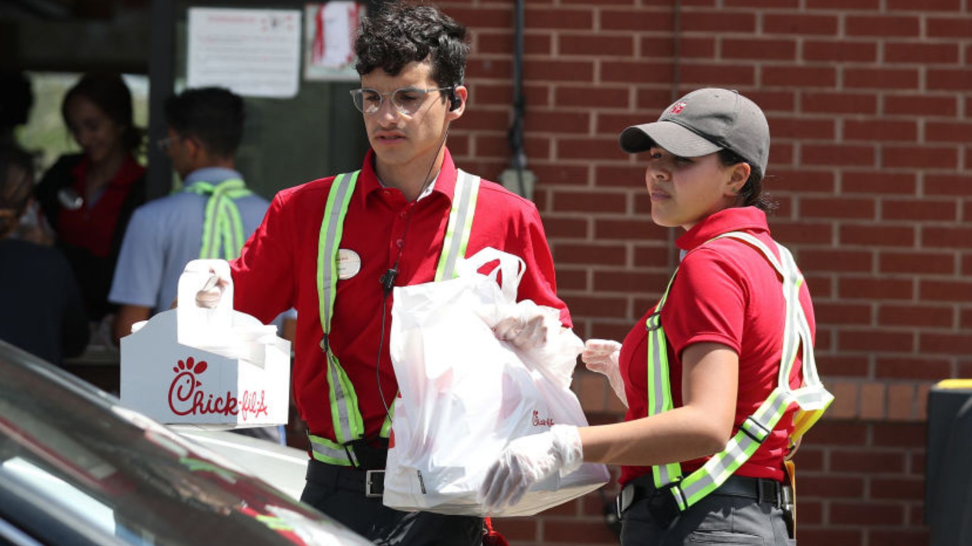 Two Chick-fil-A employees wearing red uniforms and safety vests deliver food orders to a car outside the restaurant. One holds two bags while the other holds a drink and a bag. A brick wall is visible in the background.