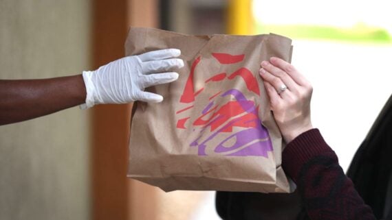 A gloved hand hands a brown paper takeout bag with a colorful logo to another person with a visible ring on their finger.