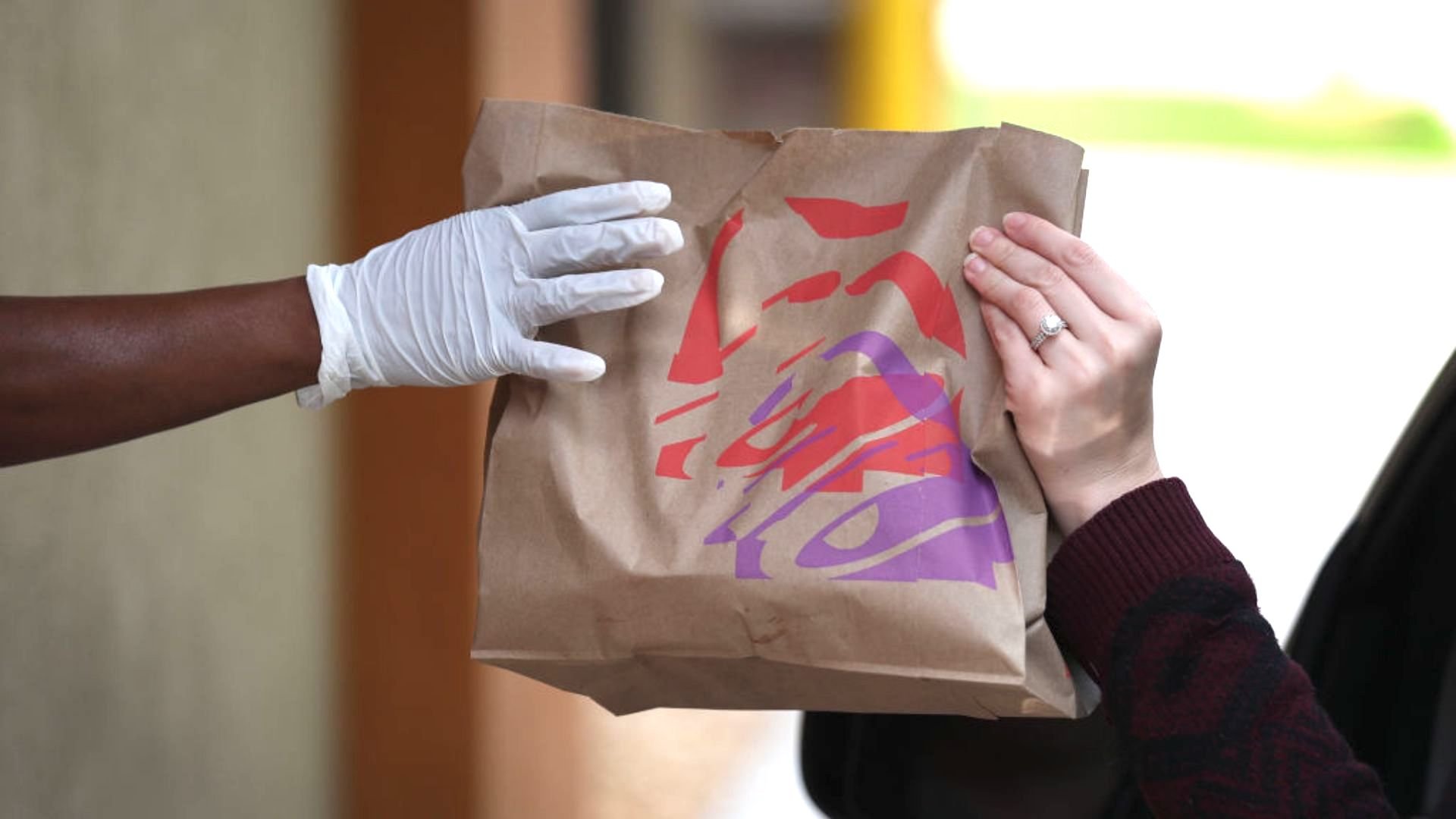 A gloved hand hands a brown paper takeout bag with a colorful logo to another person with a visible ring on their finger.