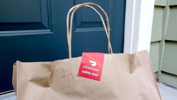 A brown paper delivery bag with a DoorDash safety seal sits in front of a dark blue door and green siding.
