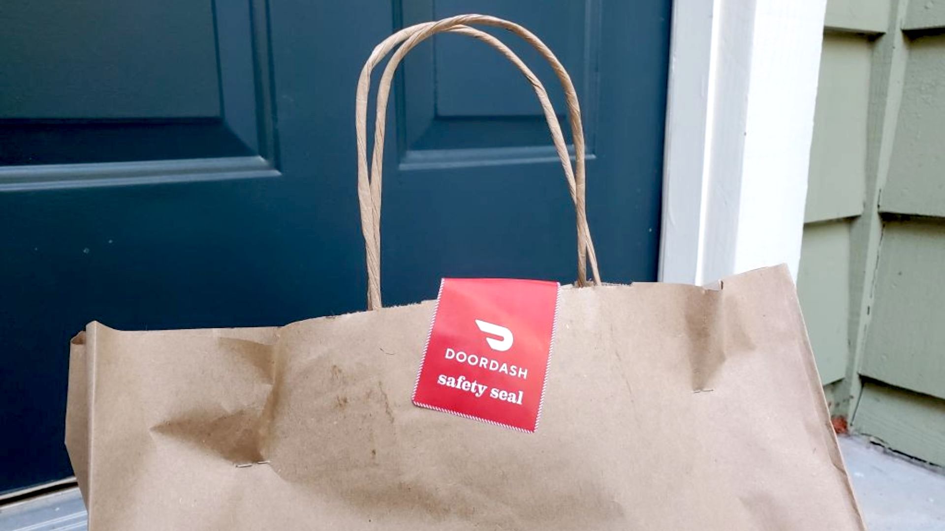 A brown paper delivery bag with a DoorDash safety seal sits in front of a dark blue door and green siding.
