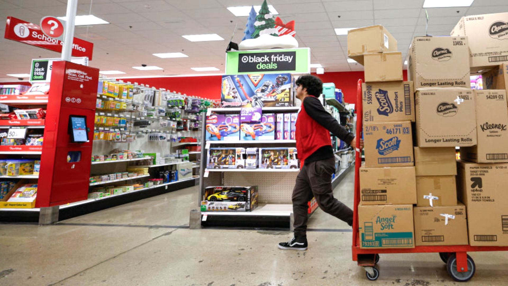 A store worker in a red vest moves a cart stacked with boxes past a Black Friday deals display in a brightly lit retail store aisle.