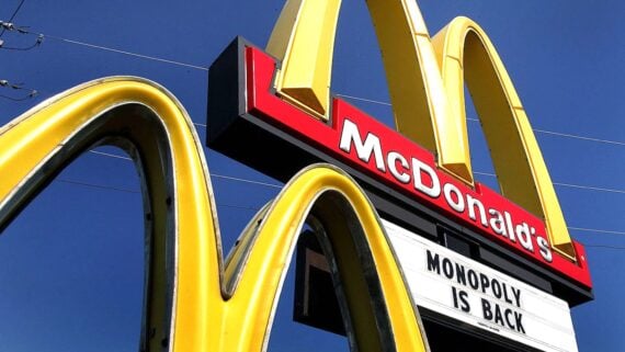Close-up of a McDonald's sign with large yellow arches and a marquee below reading "MONOPOLY IS BACK" against a clear blue sky.