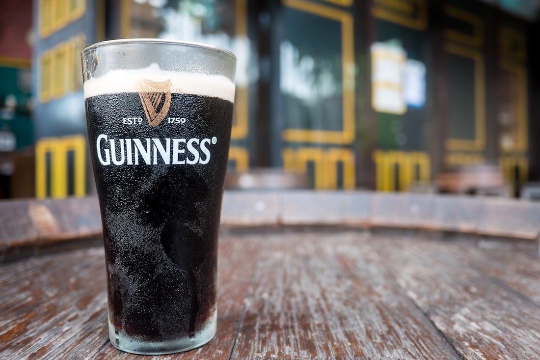 A pint glass of Guinness stout with a creamy head sits on a weathered wooden table outdoors, with a blurred background of pub-style windows and doors.