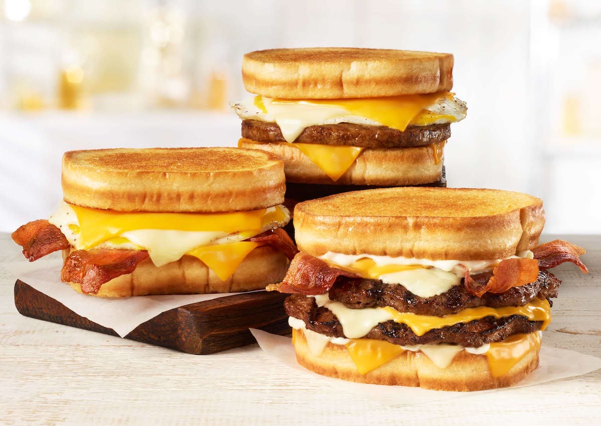 Three grilled cheese sandwiches with beef patties, melted cheese, bacon, and eggs are stacked on a light wooden surface—an irresistible addition to any new fast-food menu, showcasing crispy golden bread and layers of savory ingredients.